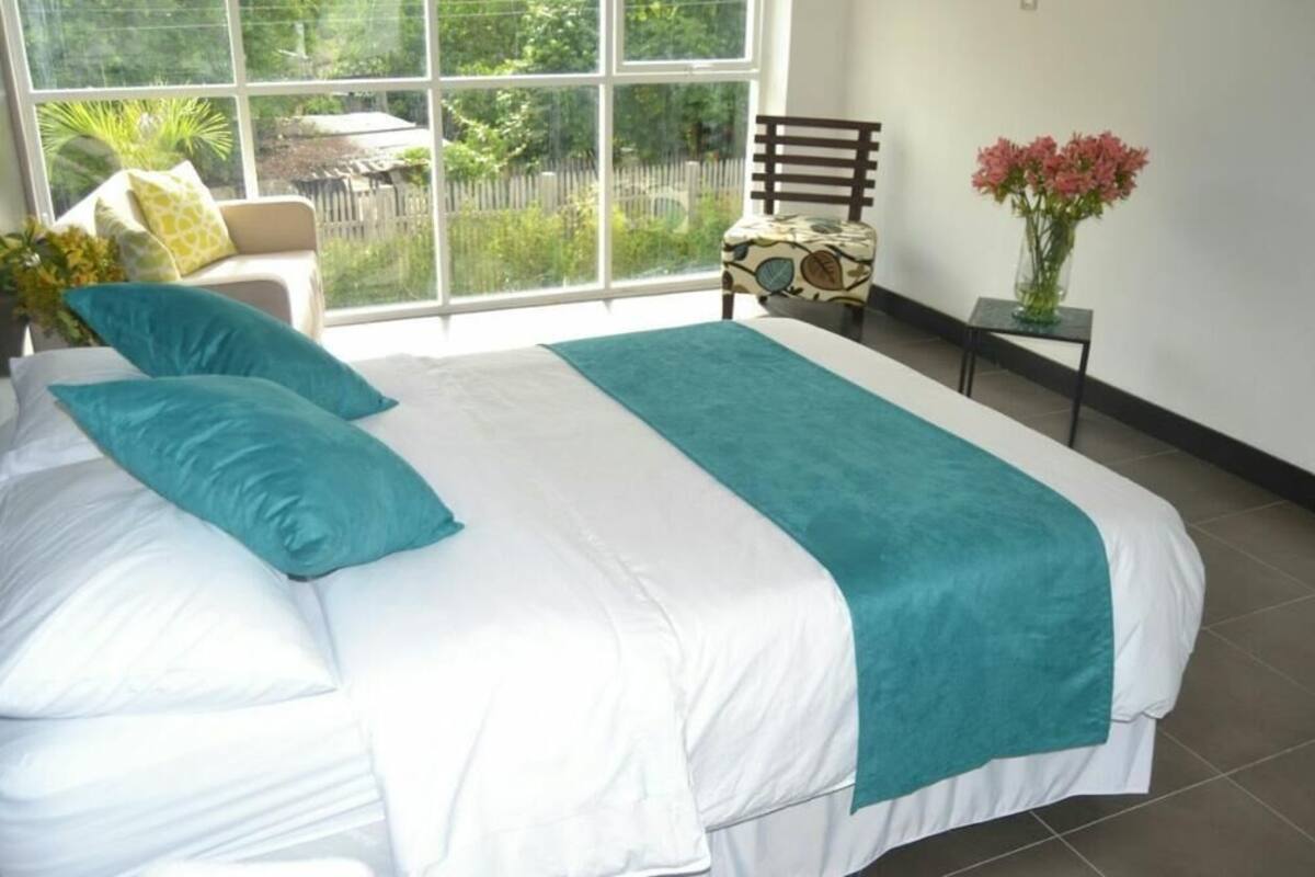 A bedroom with a large bed, a chair, a small table, and a window with a view of greenery.