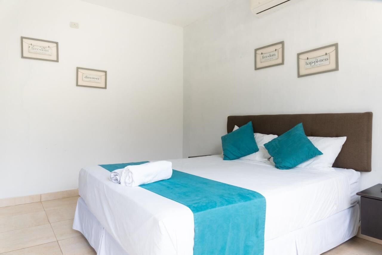 Una cama bien hecha con ropa de cama blanca y aguamarina, dos almohadas aguamarinas y una toalla doblada al lado.