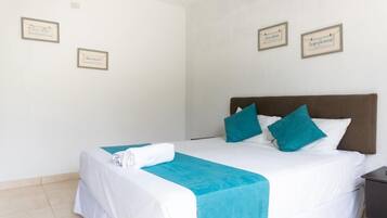 Una cama bien hecha con ropa de cama blanca y aguamarina, dos almohadas aguamarinas y una toalla doblada al lado.