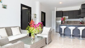Una sala moderna con un sofá, una mesa de centro de cristal y un jarrón de flores. Se observa una zona de cocina con armarios oscuros y un taburete de bar.