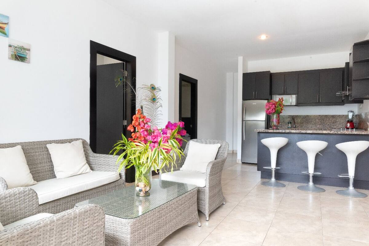 A modern living room with a sofa, a glass coffee table, and a vase of flowers. There is a kitchen area with dark cabinets and a bar stool.