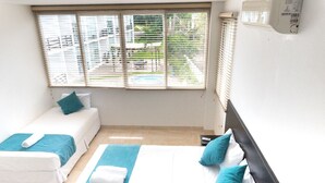 Habitación de hotel con dos camas, un ventanal con persianas, un ventilador de techo y vistas a una piscina y zonas verdes.