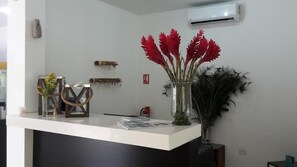 Un mostrador de recepción con una superficie blanca, un jarrón con flores rojas, un marco de madera decorativo y un aire acondicionado instalado en la pared.