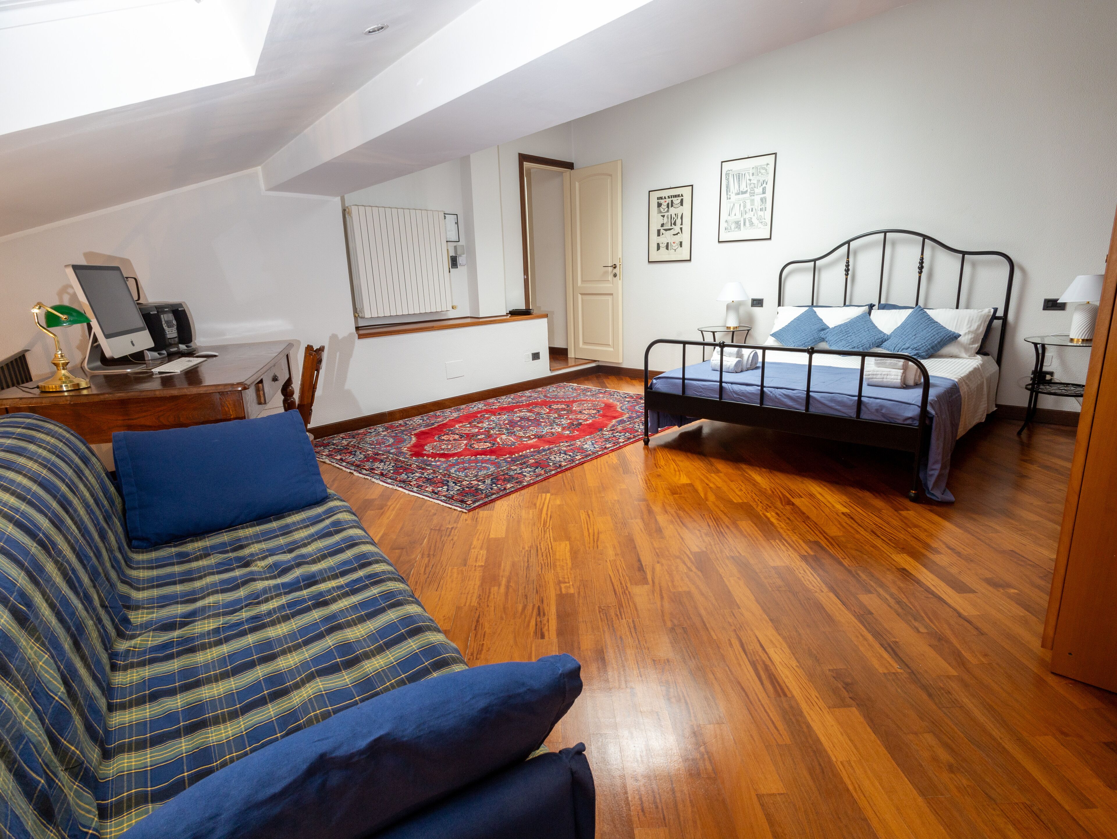 A bedroom with a bed, a desk with a computer, a sofa, and a red rug.