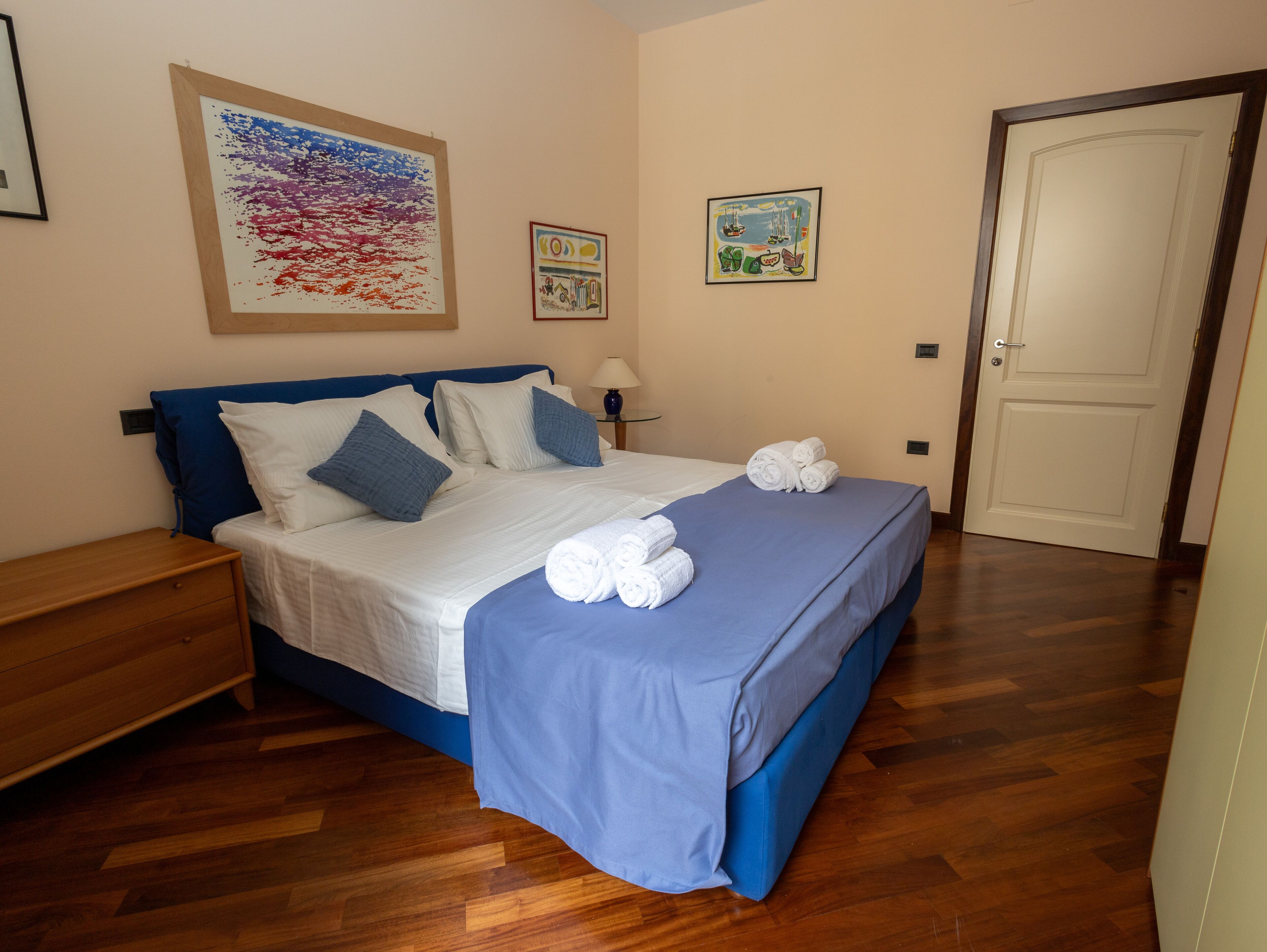 A neatly made bed with blue bedding and white pillows, a wooden nightstand, and a door to the right.