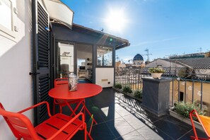 Un balcon avec une table et des chaises rouges, un pot de fleurs et une vue sur un bâtiment surmonté d’un dôme.