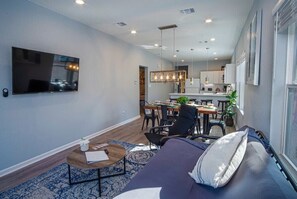 Una sala de estar moderna con un sofá, una mesa de centro y un televisor. Hay un comedor con una mesa y sillas, y una cocina al fondo.