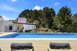 A poolside area with lounge chairs and a table, surrounded by a white building and trees. - Villa Can Rafalet - Five Bedroom Villa, Sleeps 10 (Ibiza)