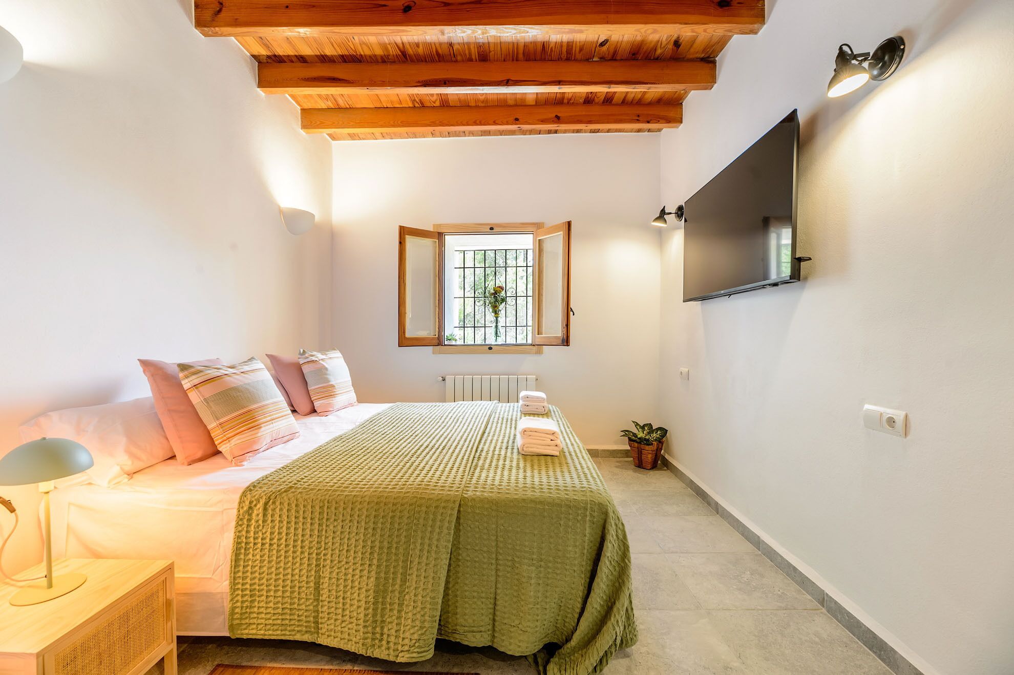 Un dormitorio con cama, mesita de noche, lámpara, ventana y un televisor de pantalla plana montado en la pared.