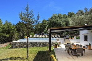 Un área junto a la piscina con una zona de estar techada, una mesa de comedor preparada para cuatro personas y una pared de piedra.