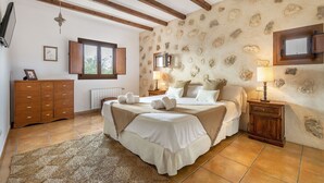 A bedroom with a stone wall, wooden beams, a bed with white linens, a wooden dresser, and a window with shutters. - New! Villa Can Jaume Arabí de Baix (Puig d'en Valls)