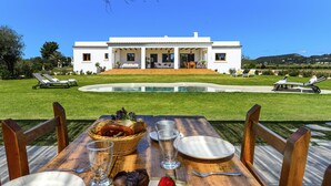 A dining table with plates, cutlery, and a basket of bread and pastries, set against a backdrop of a white villa with a pool and garden furniture. - New! Villa Can Jaume Arabí de Baix (Puig d'en Valls)