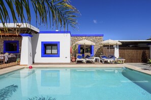 Un área de piscina con sillones de descanso y sombrillas, un muro de piedra y un edificio azul y blanco con una ventana y una puerta.