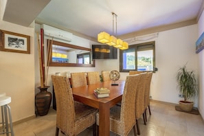 A dining area with a wooden table and woven chairs, a TV mounted on the wall, and a large window with a view of greenery. - New! Villa Cas Pla 33 (Ibiza)