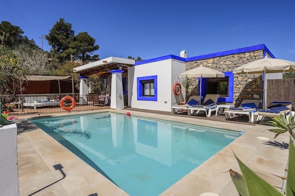 A swimming pool with lounge chairs and umbrellas, surrounded by a stone wall and a white building with blue accents. - New! Villa Cas Pla 33 (Ibiza)