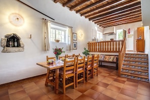 Un comedor con una mesa y sillas de madera, un estante de piedra con botellas, y una ventana con cortinas.