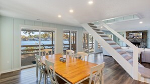 Un comedor espacioso con una mesa de madera y sillas blancas, una escalera que conduce a una sala de estar y ventanas amplias con vistas al exterior.