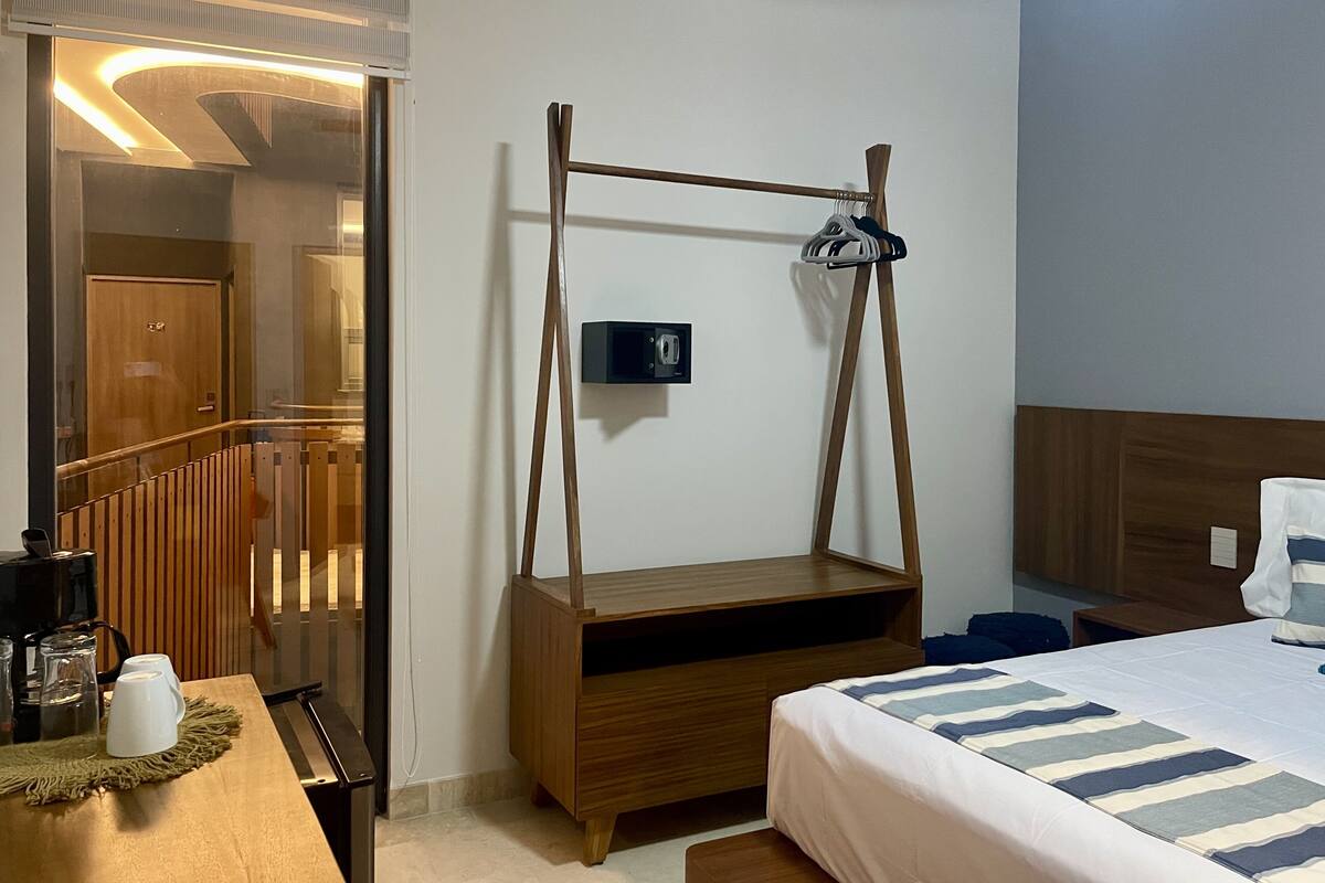 Habitación de hotel con cama de madera, mesita de noche con cafetera, sillón colgante y vista al pasillo.