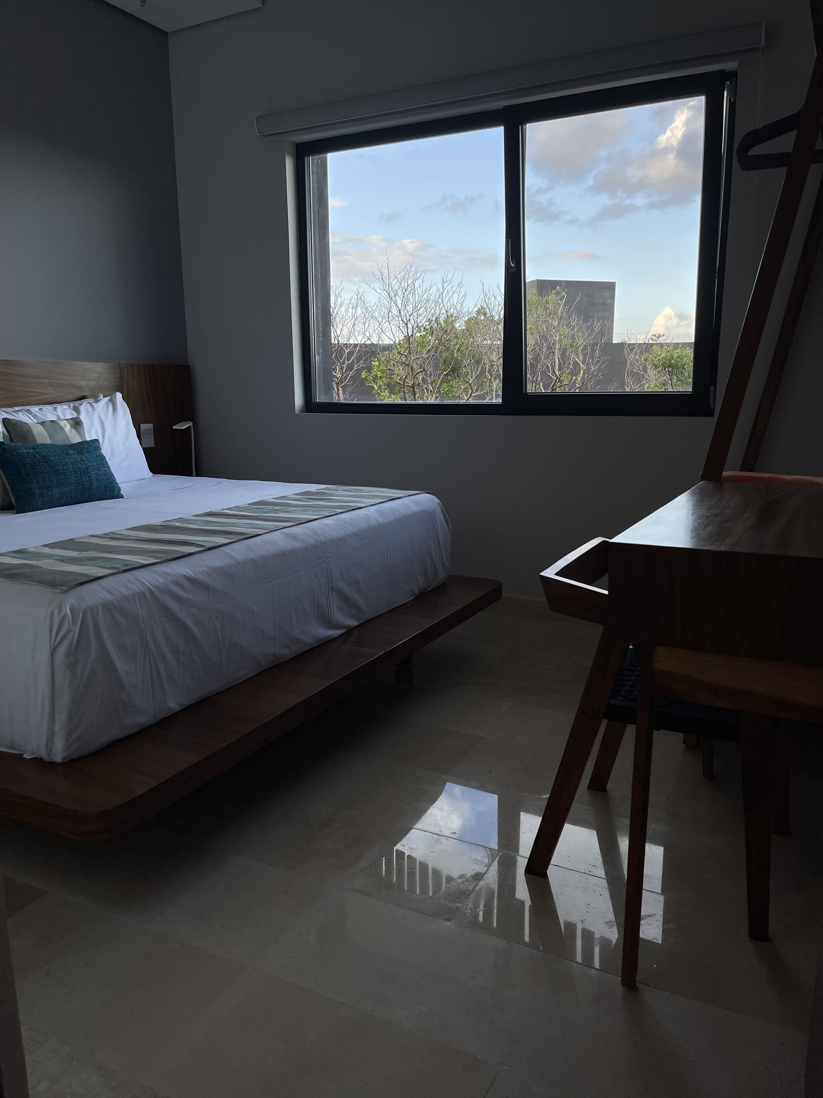 A bedroom with a bed, a desk, and a window with a view of trees and buildings.