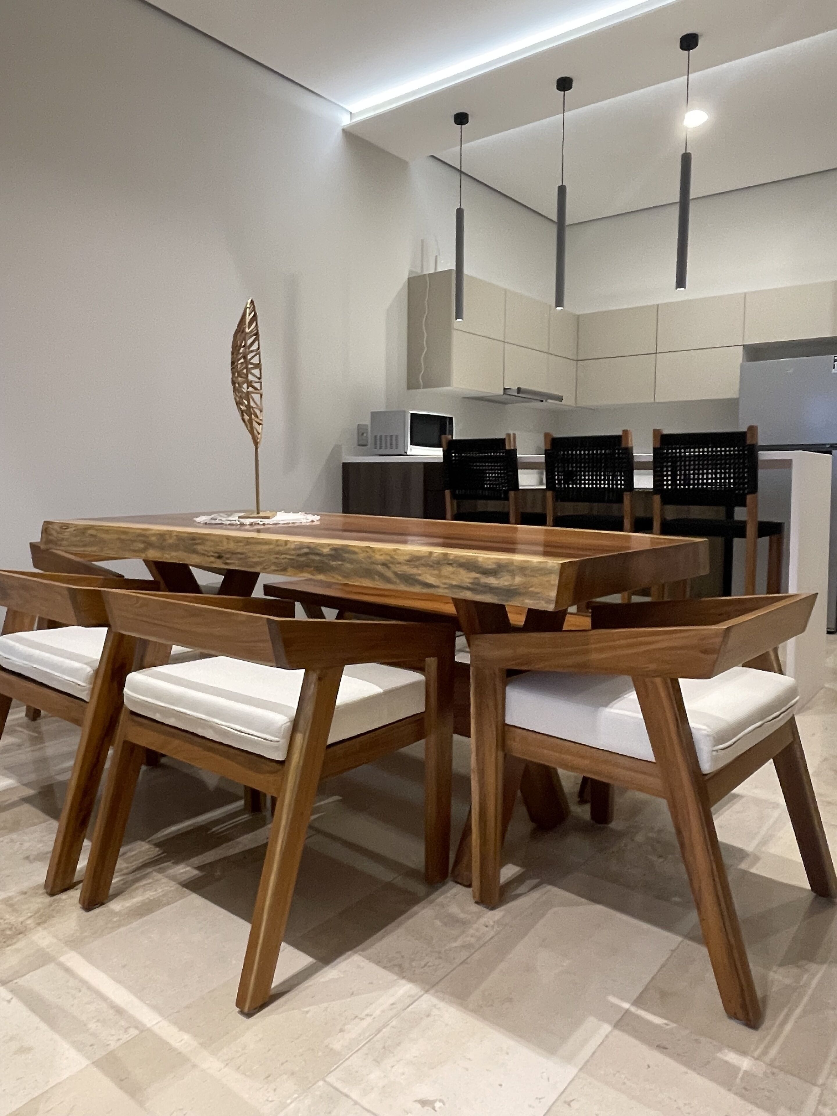 A modern dining area with a wooden table and chairs, a decorative leaf sculpture, and a kitchen in the background.