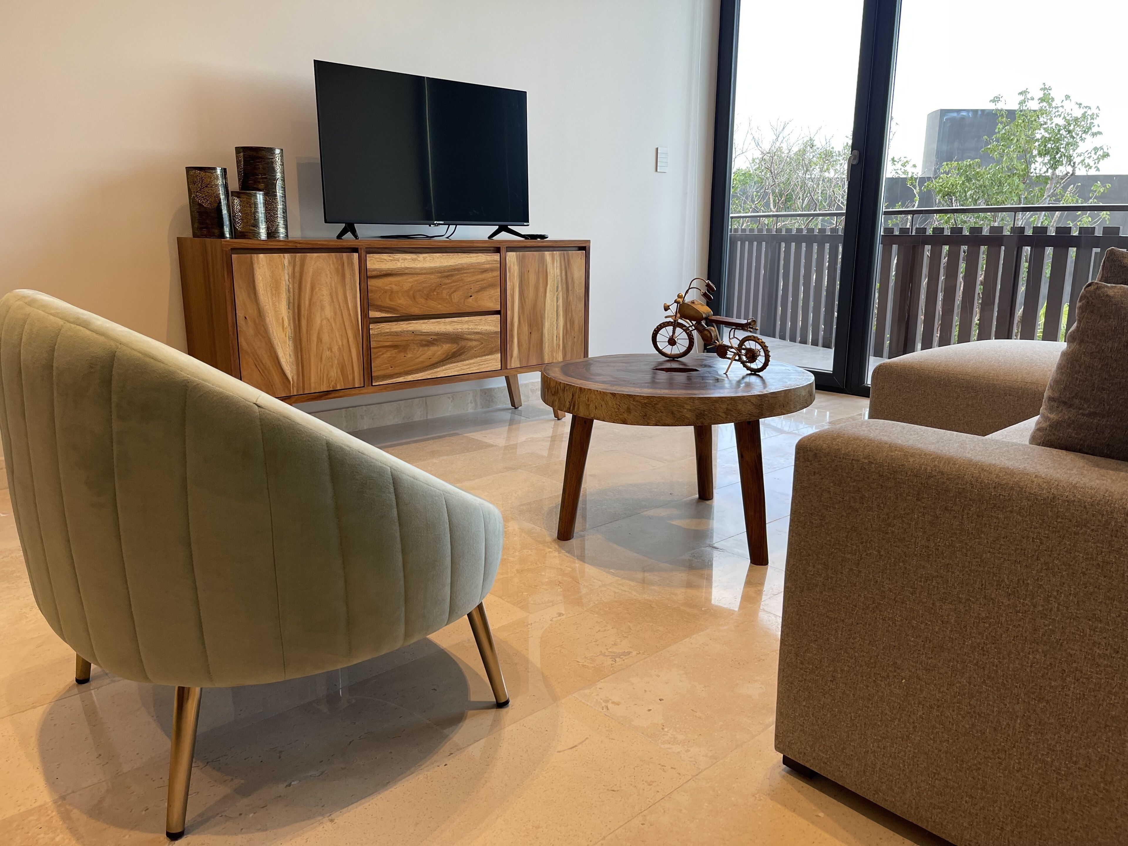 A modern living room with a sofa, armchair, and a small round table with a decorative item. A television is mounted on a wooden cabinet, and a balcony with a view of trees and buildings is visible through large windows.