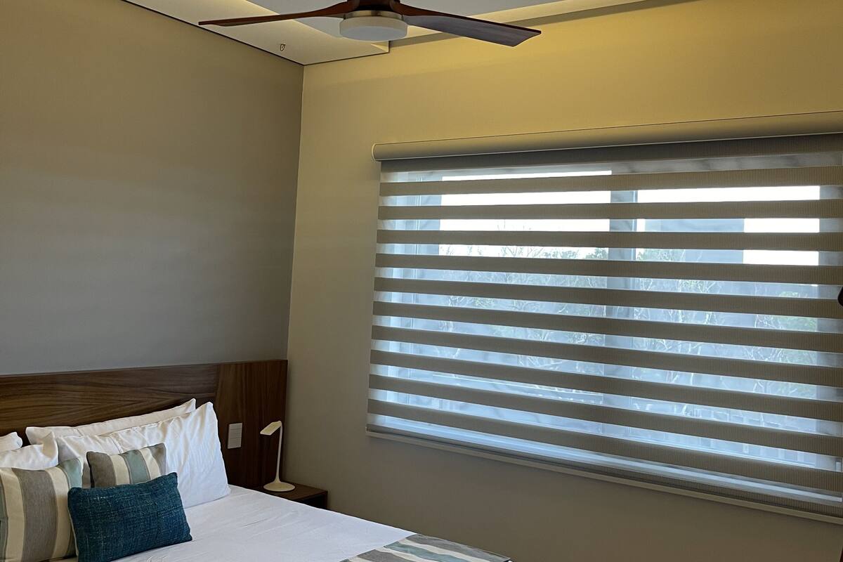Un dormitorio con cama, ventilador de techo, ventana con persianas y mesita de noche.