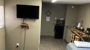 A room with a bed, a desk with a chair, a television mounted on the wall, and a kitchenette area.