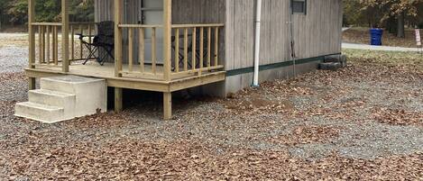 A small wooden cabin with a porch and a covered area, surrounded by gravel and trees.