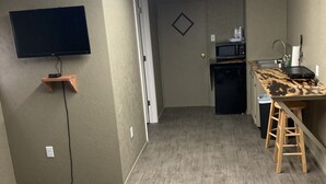 A compact living space with a flat-screen TV mounted on the wall, a kitchenette with a microwave and sink, and a small wooden stool.