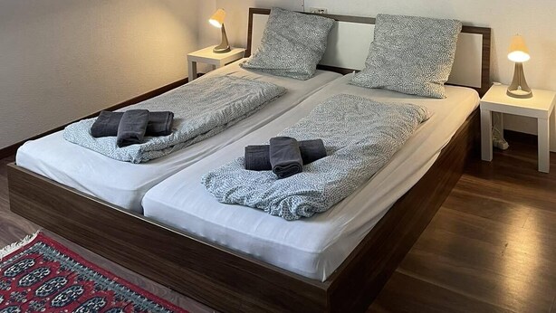 A bedroom with a double bed, two bedside tables with lamps, a large landscape painting on the wall, and a patterned rug. - Apartment Beny (Bad Bellingen)