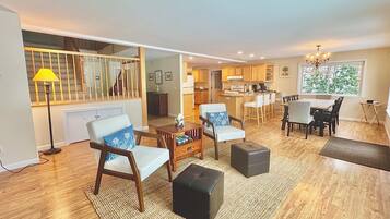 Un vaste salon comprenant un canapé, une table basse et un pouf, un coin repas avec des chaises et une table, une cuisine avec des meubles en bois, et un escalier menant à l’étage supérieur.