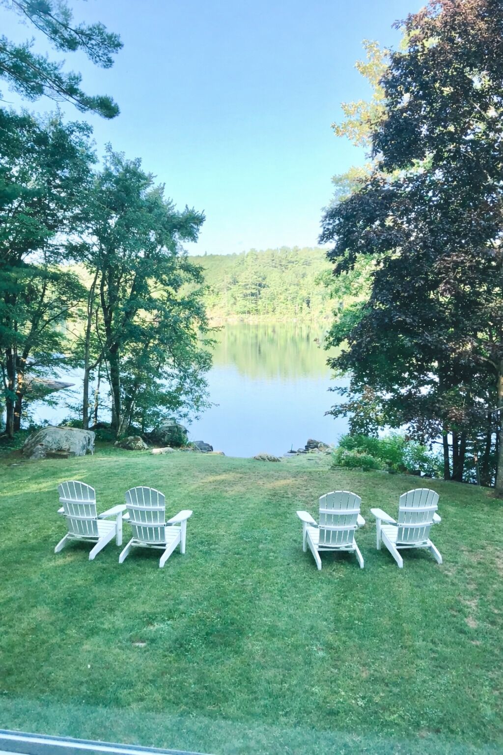 Une zone herbeuse avec quatre fauteuils Adirondack disposés face à un lac paisible, entouré d’arbres.