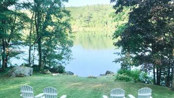 A grassy area with four Adirondack chairs facing a calm lake surrounded by trees.