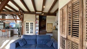 Ein großzügiger Wohnbereich mit einem blauen Sofa, Holzbalken, einer Küche im Hintergrund und einer Treppe, die nach oben führt.