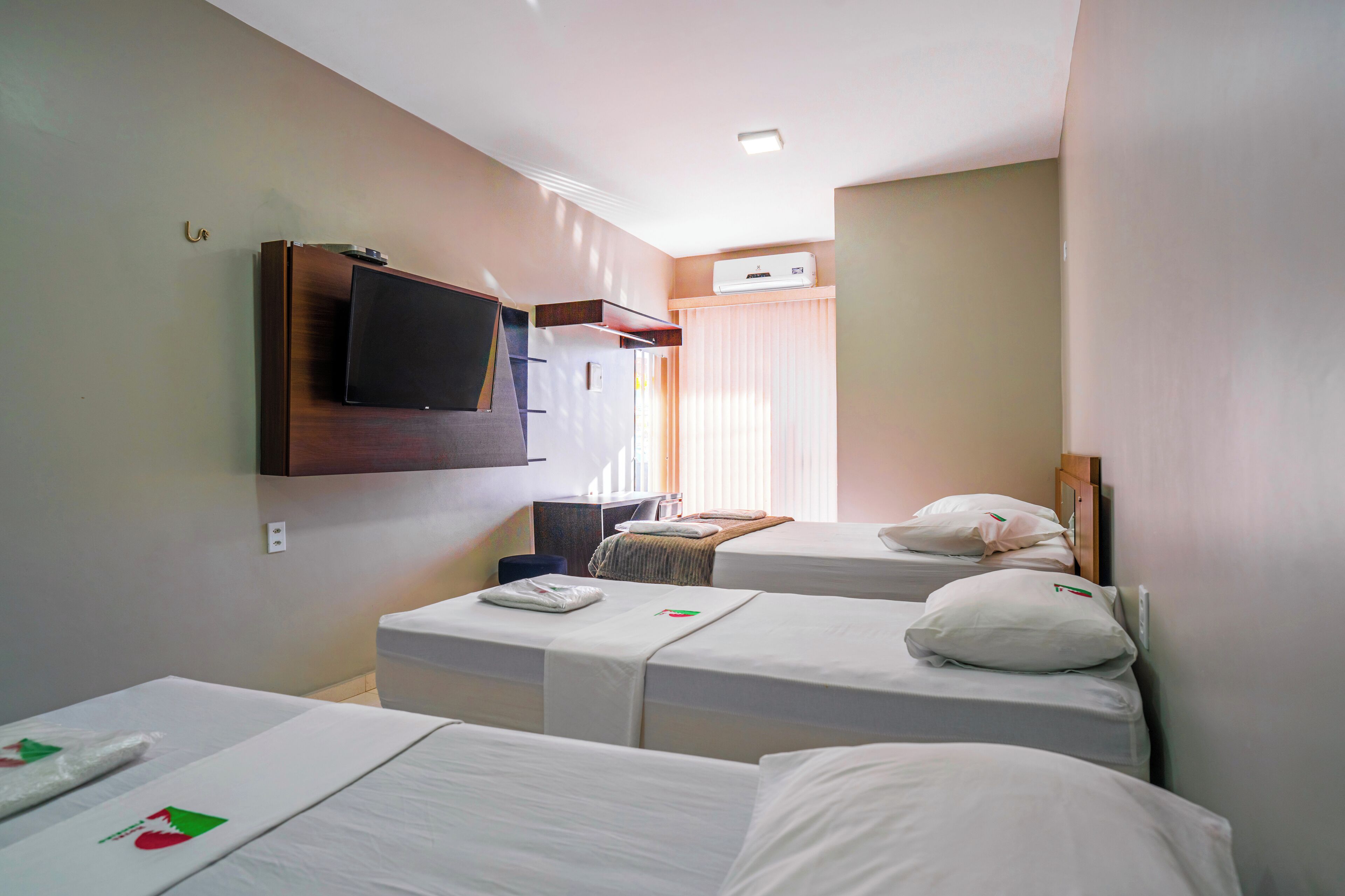 A hotel room with two single beds, a flat-screen TV mounted on the wall, an air conditioning unit, and a small desk with a chair.