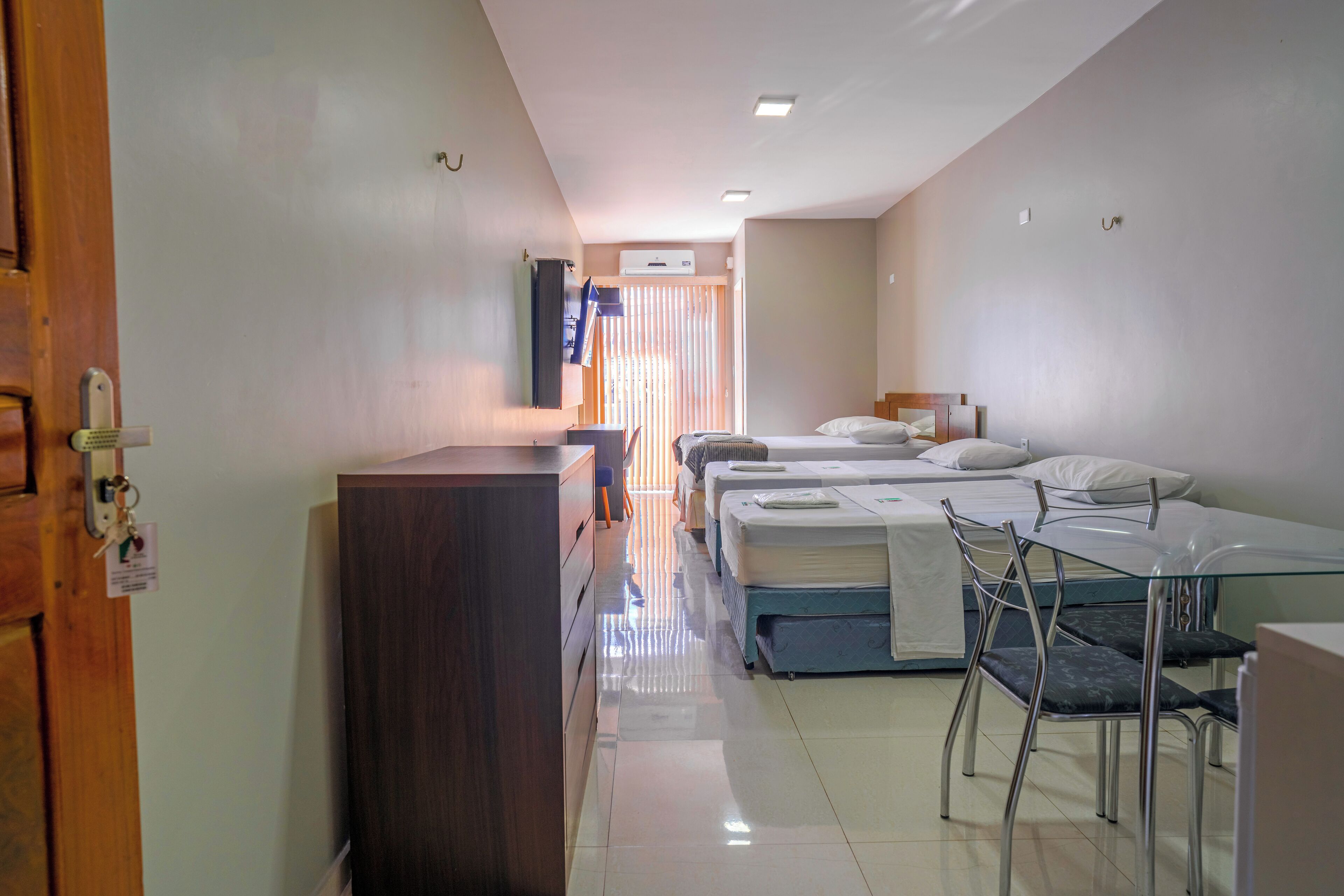 Habitación de hotel con un escritorio de madera, una mesa de vidrio, tres camas y un baño con ducha.