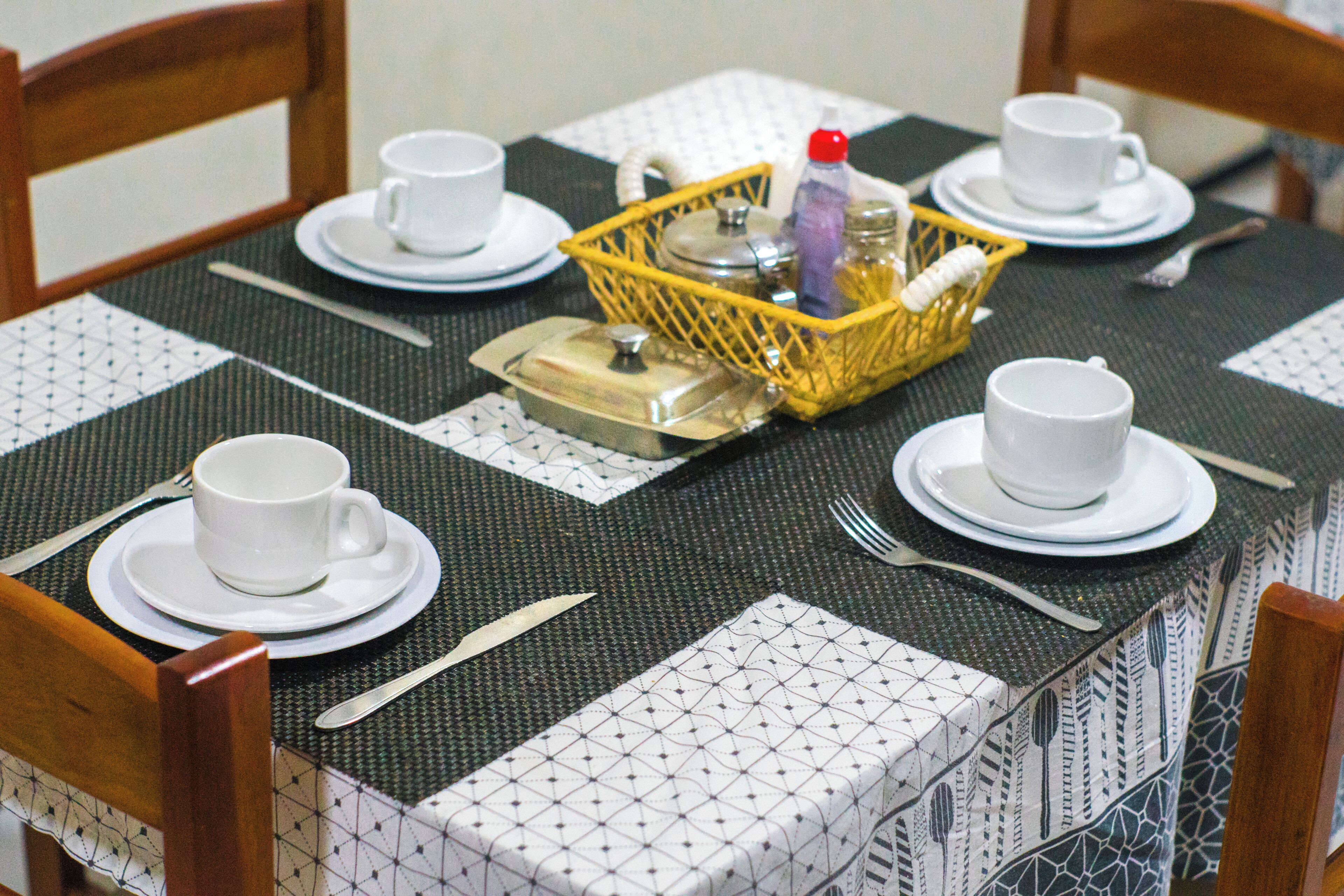 Una mesa de comedor dispuesta con tazas, platillos y cubiertos blancos, una cesta amarilla con condimentos y un mantel estampado.