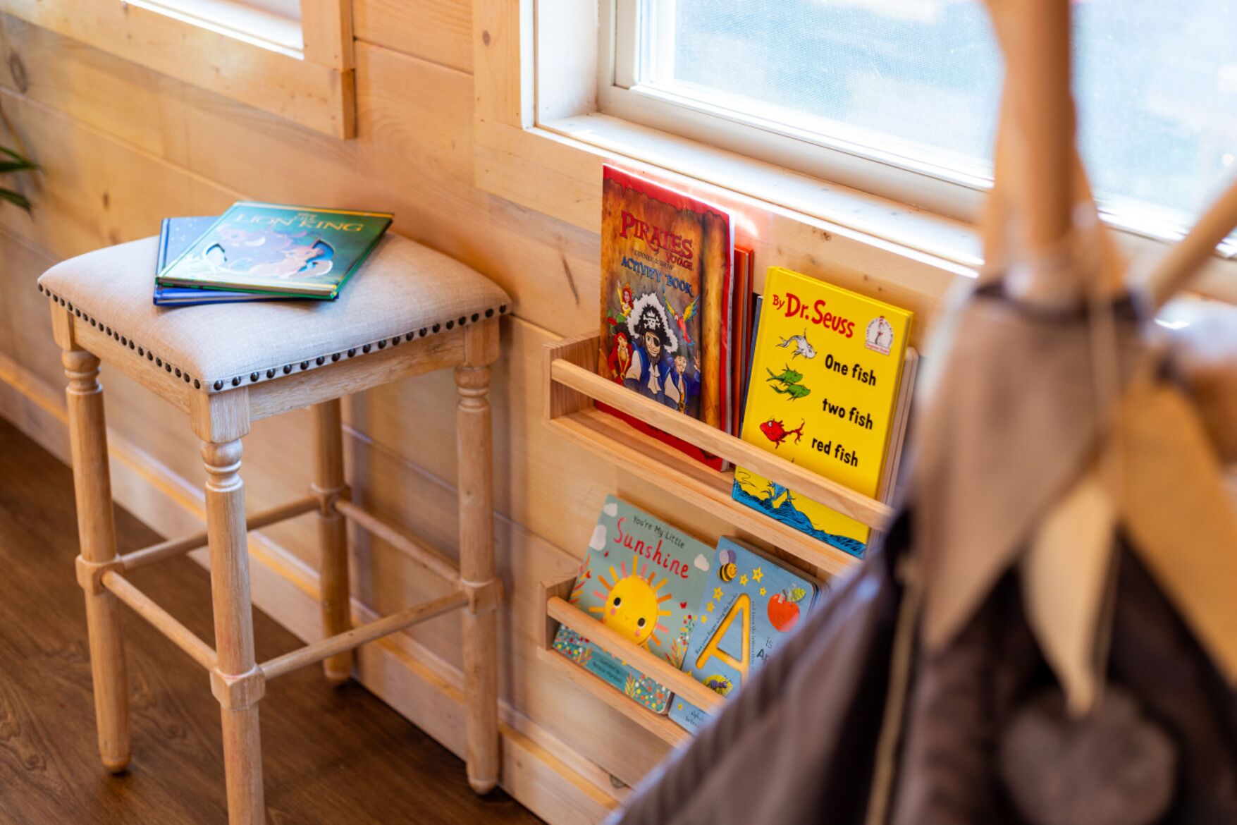 A wooden chair with a cushion, a bookshelf with children's books, and a window with natural light.