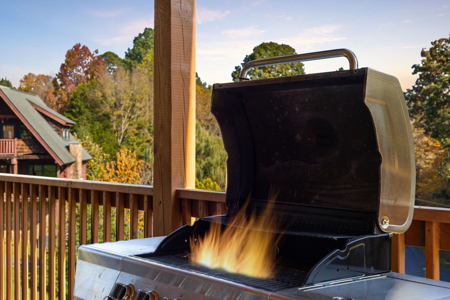 A barbecue grill with flames, set on a wooden deck with a house in the background.
