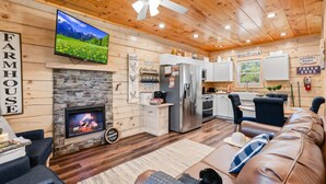 A cozy farmhouse interior with a stone fireplace, a television mounted above it, a kitchen area with a refrigerator and oven, and a leather sofa. - Enchanted Forest Hideaway by Ghosal Luxury Lodging (Sevierville)