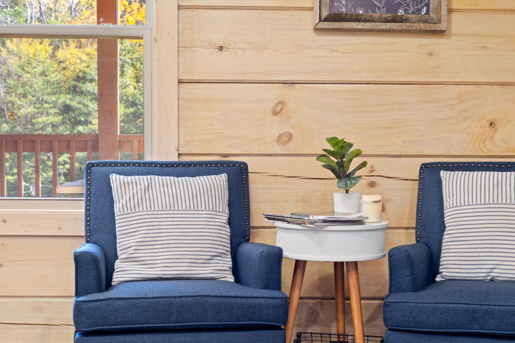 Two blue armchairs with striped pillows, a small white table with a plant, and a framed picture on a wooden wall.