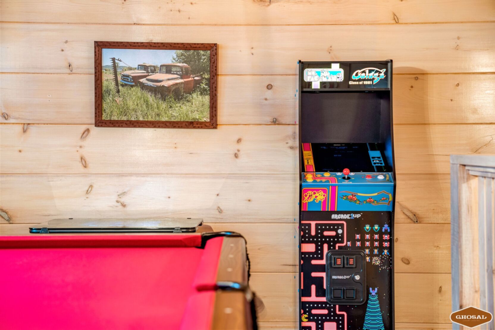 Un mur en bois orné d’un tableau représentant de vieilles voitures, une borne d’arcade aux couleurs vives et une table rouge.
