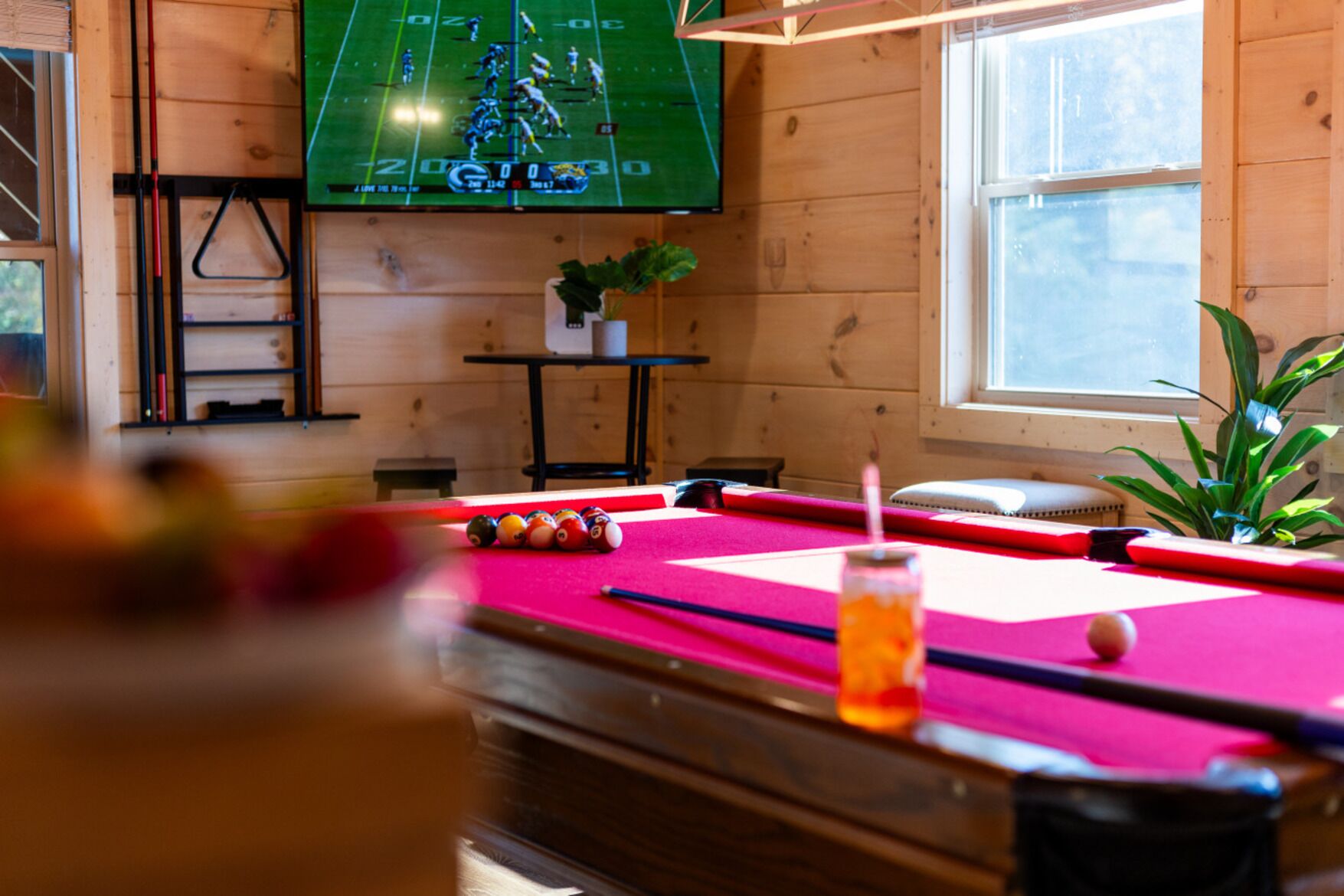 A pool table with balls, a television displaying a sports game, and a window with natural light.
