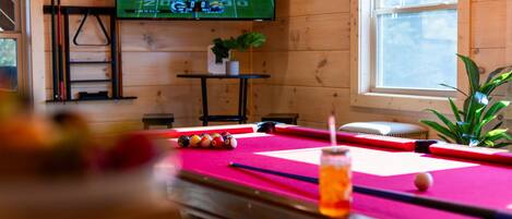 A pool table with balls, a television displaying a sports game, and a window with natural light.