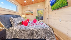 A bedroom with a patterned bedspread, a ceiling fan, a painting, and a TV on the wall. - Enchanted Forest Hideaway by Ghosal Luxury Lodging (Sevierville)