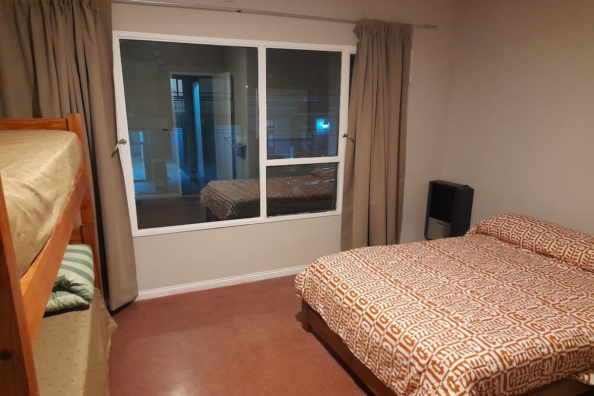 Ein Schlafzimmer mit einem Etagenbett, einem Einzelbett mit gemusterten Textilien, einem Fenster mit Vorhängen und Blick auf einen Balkon.