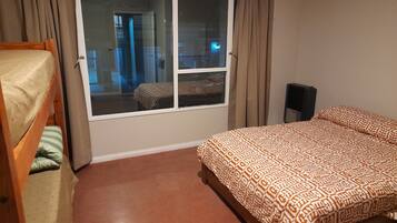 A bedroom with a bunk bed, a single bed with patterned bedding, a window with curtains, and a view of a balcony.