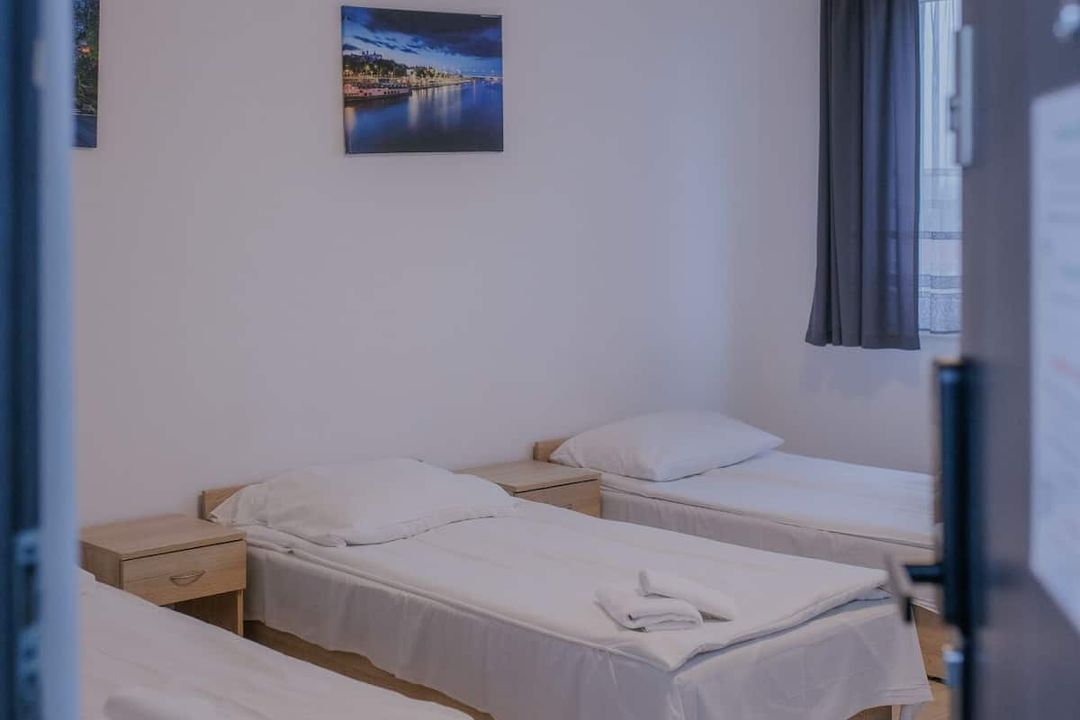 Une chambre avec trois lits simples, une table de chevet, un tableau accroché au mur et une fenêtre avec des rideaux.