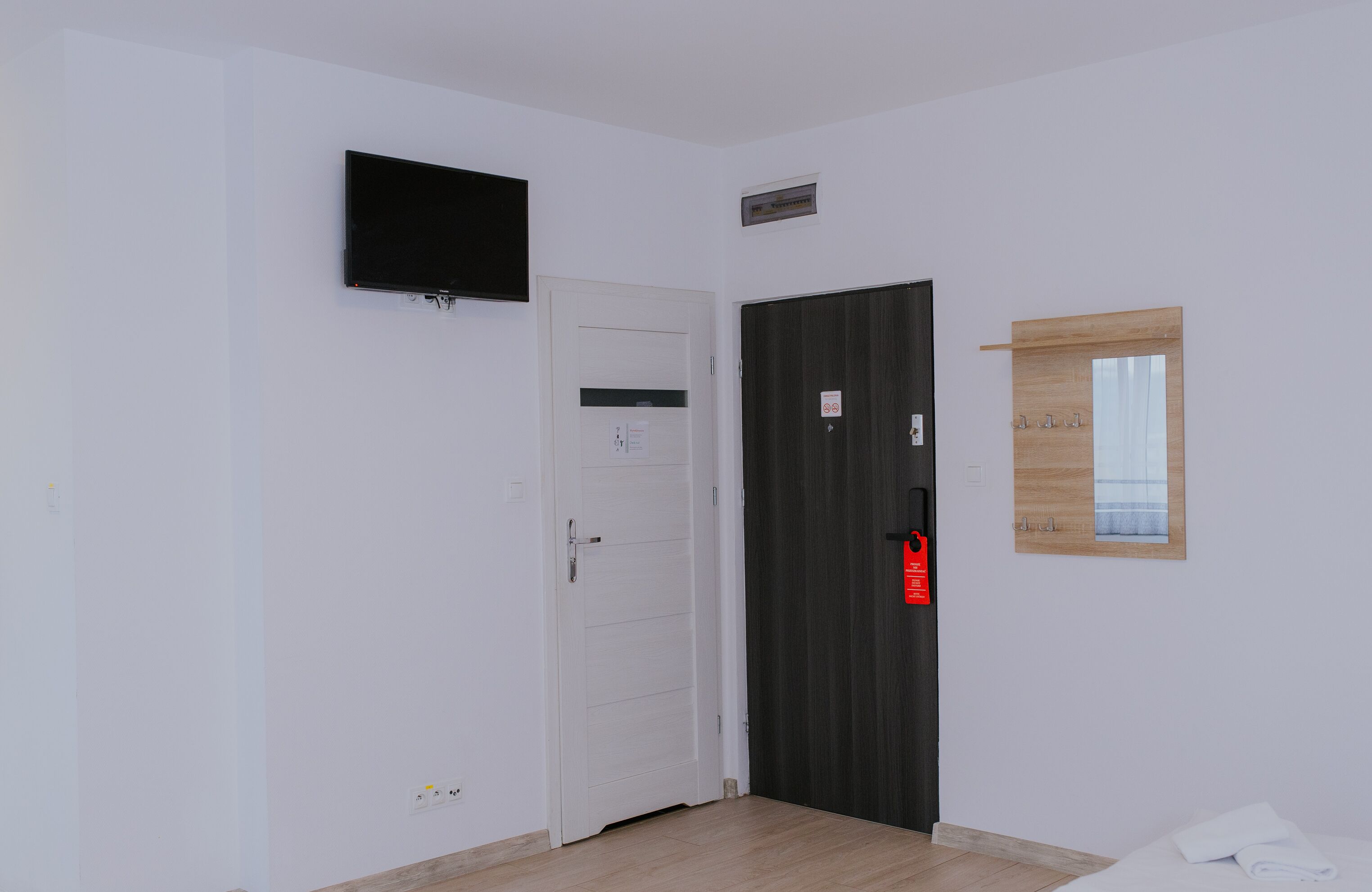 A room with a white door, a black door, a mounted TV, a small window with a wooden frame, and a white towel on the floor.