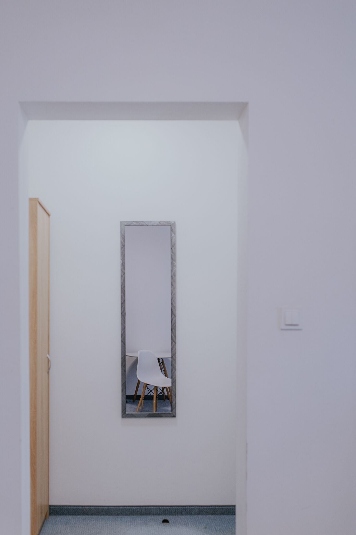 A minimalist room with a large mirror on the wall, a wooden door, and a small white chair.